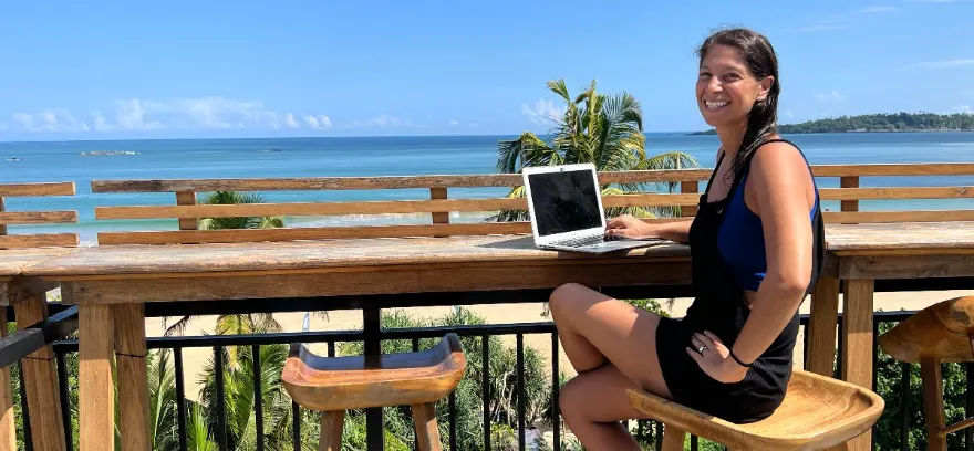 A woman smiling at a laptop on a beachfront deck overlooking the ocean in Bali, Indonesia