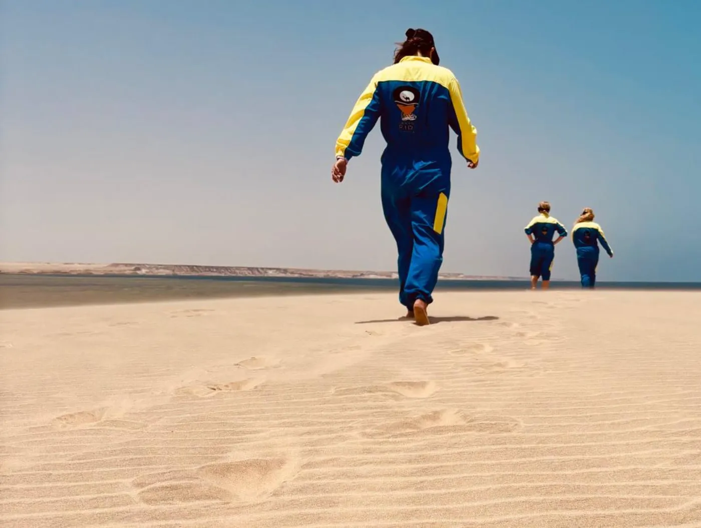 Three people in blue-and-yellow jumpsuits walking across a sand spit toward the sea in Dakhla, Morocco