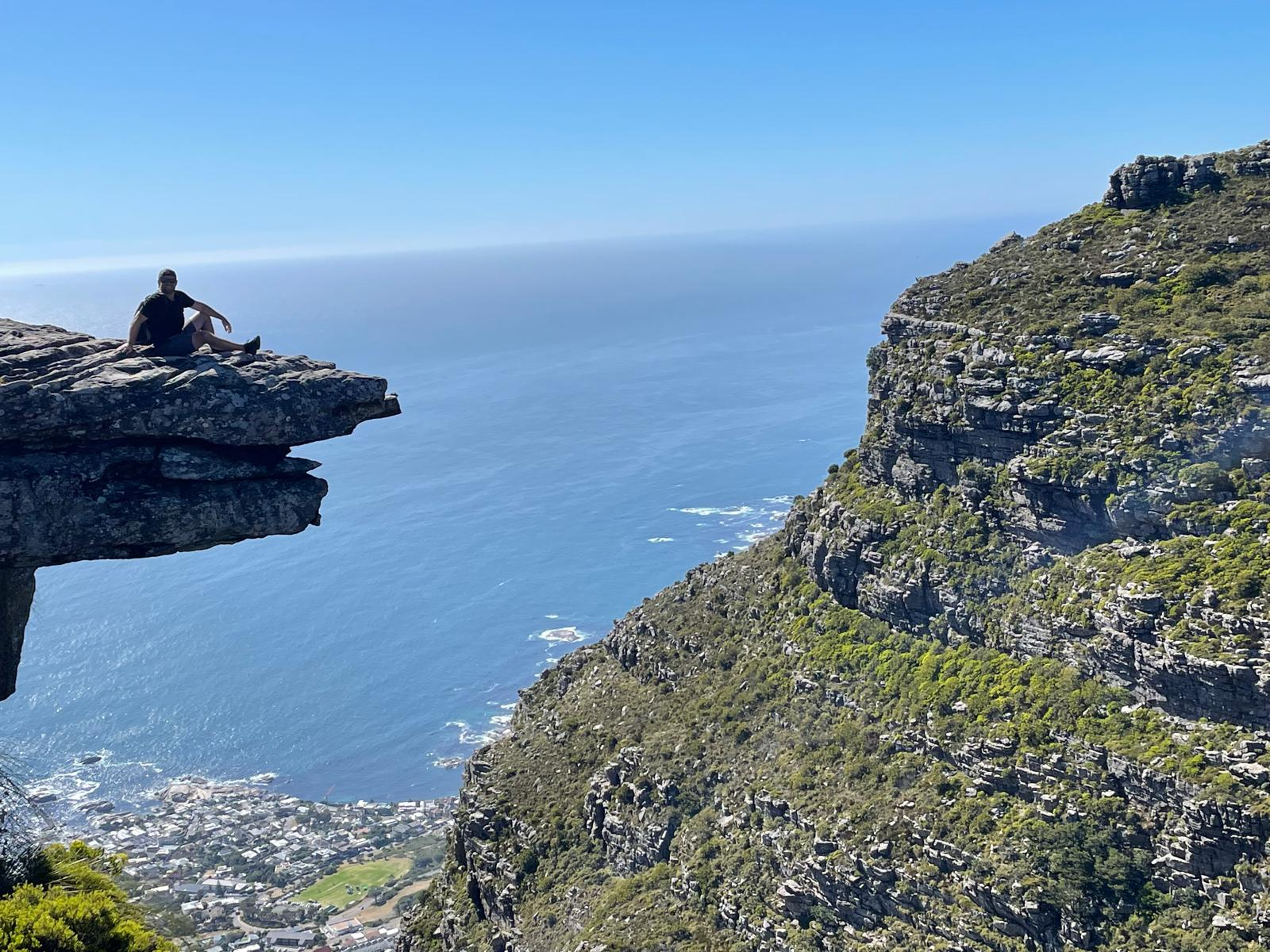 A hiker sitting on a rock ledge high above the ocean and town on Table Mountain, Cape Town